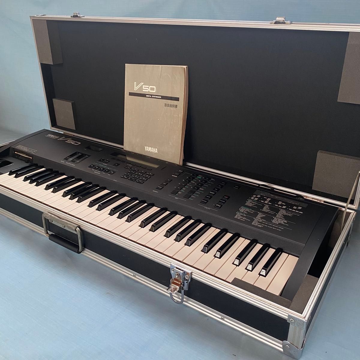 YAMAHA V50 synthesizer inside an open hard case with the V50 instruction manual propped against the lid.