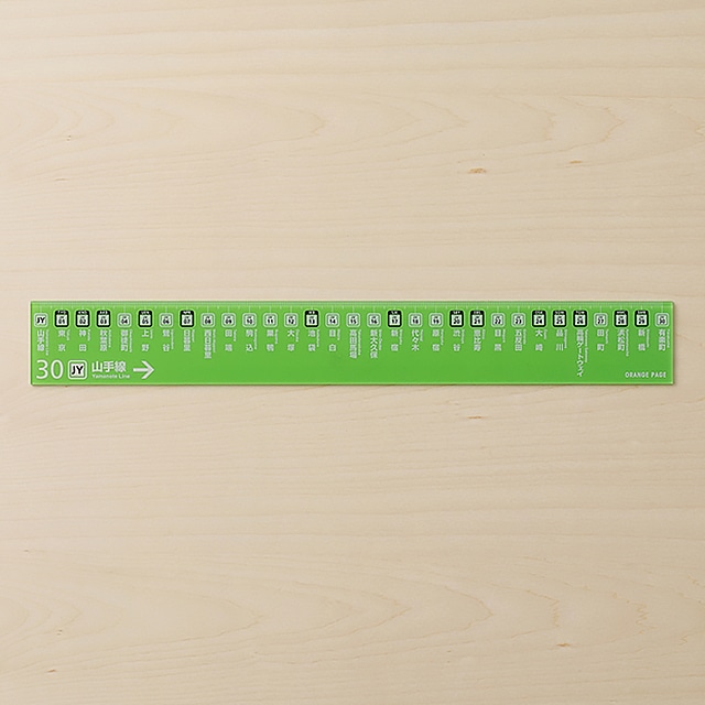 A green plastic 30 cm ruler printed with Yamanote Line station names and icons on a light wood surface.