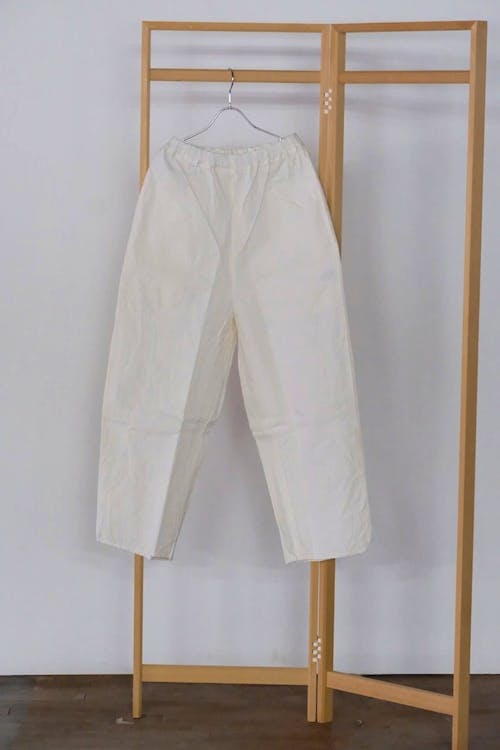 White YAMMA MAN men's cotton balloon pants hanging on a hanger from a wooden clothing rack against a plain wall.