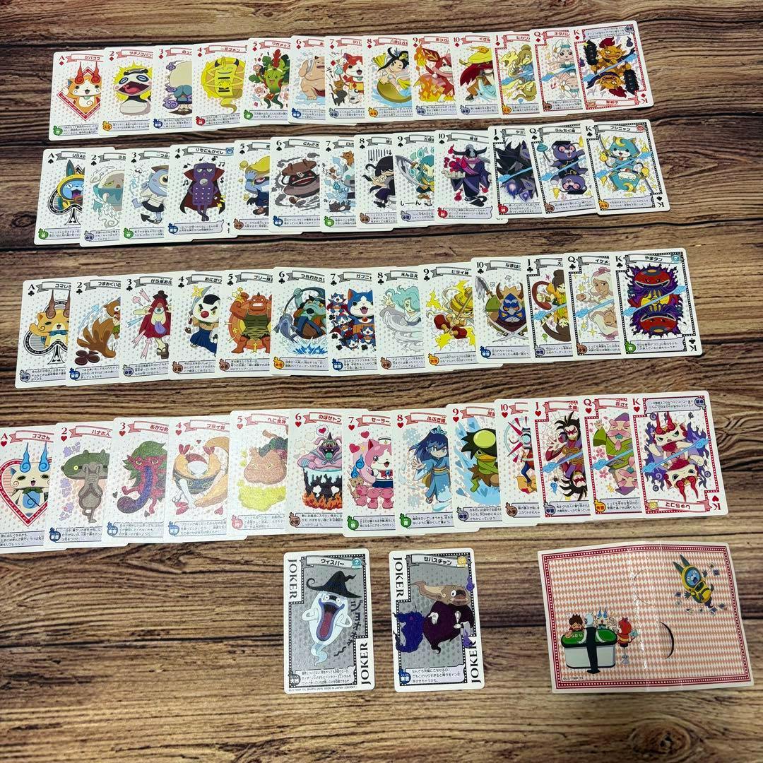 Yo-kai Watch themed playing cards spread out on a wooden surface showing many illustrated card faces, two jokers, and a card back.