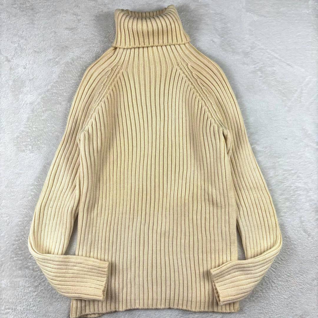 Beige ribbed turtleneck wool sweater laid flat showing back, sleeves, and folded collar on a light background.