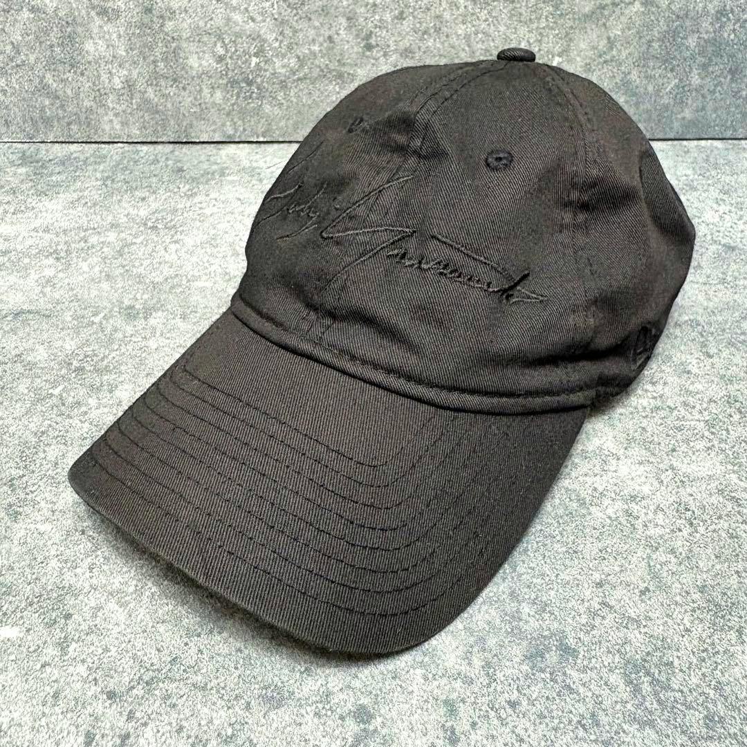 Black Yohji Yamamoto New Era cap with tonal embroidered signature on the front, photographed at an angle on a grey surface.