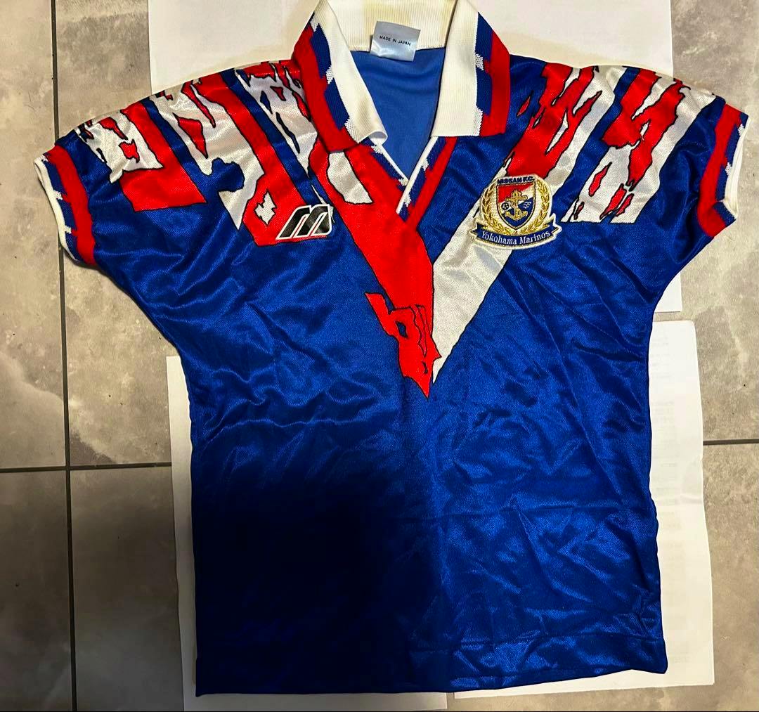Blue Yokohama Marinos Mizuno uniform (front) laid flat showing the club crest, Mizuno logo, and red-white accents.