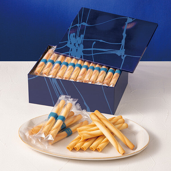 Blue decorative box of individually wrapped cigar-shaped cookies with several cookies arranged on a white plate.