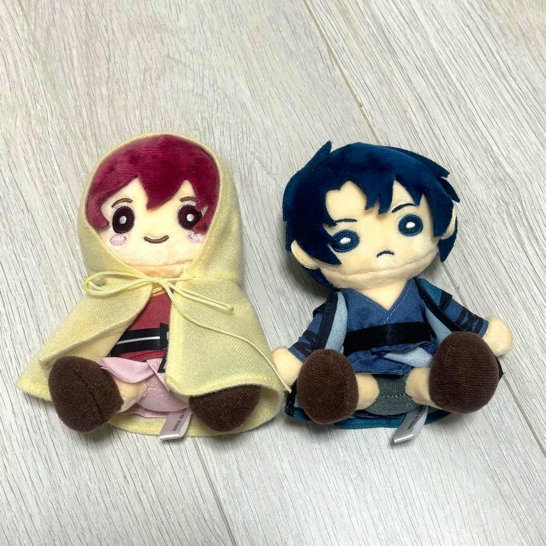 Front view of two mascot plush toys from 暁のヨナ, one in a yellow hooded cape and one with blue hair and a blue outfit on a light wood floor.