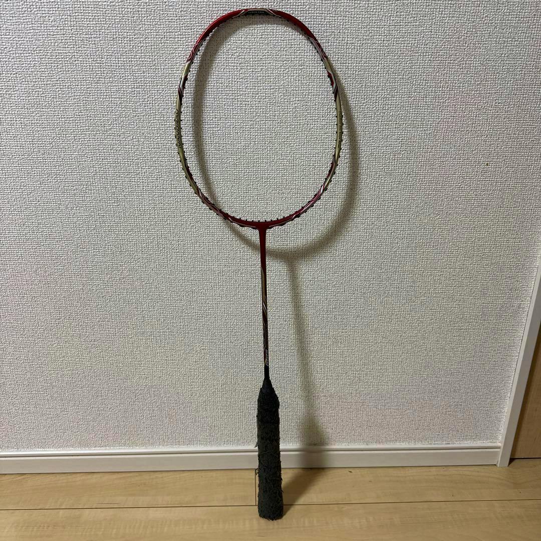 Full-length YONEX badminton racket leaning against a wall on a wooden floor.