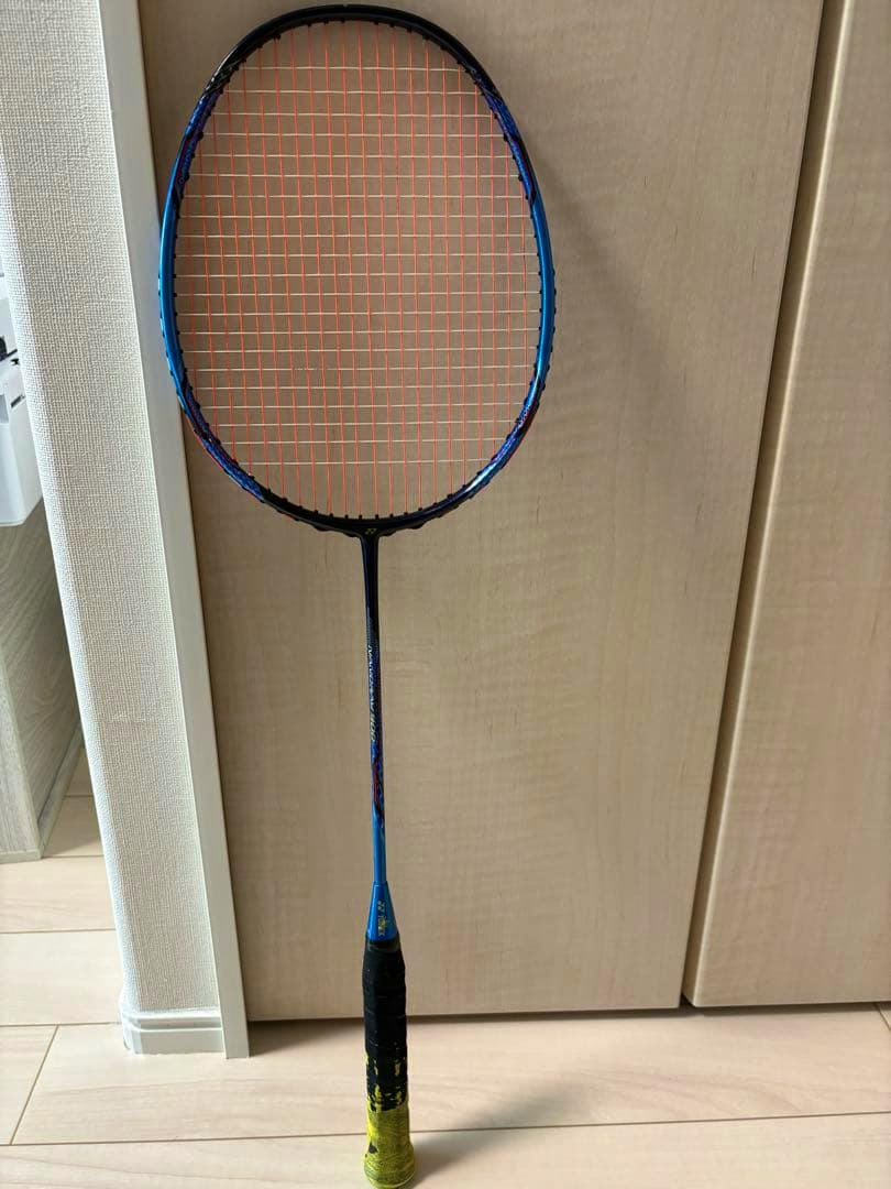 Full Yonex Nanoray900 badminton racket standing upright against a wooden background, showing a blue frame, orange strings, and a black-and-yellow grip.