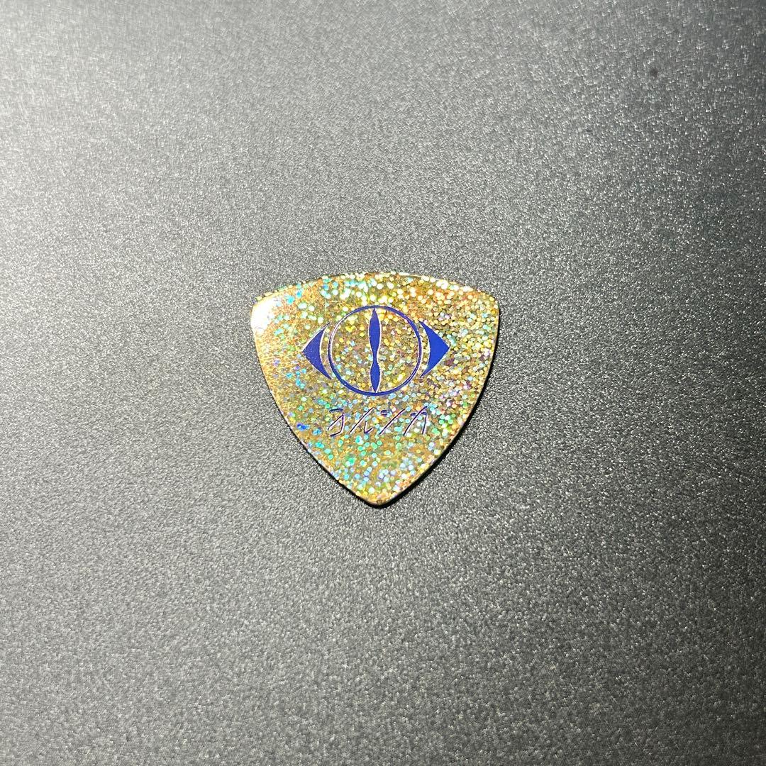 Gold glitter triangular guitar pick with a blue eye logo and small printed text on a dark background.
