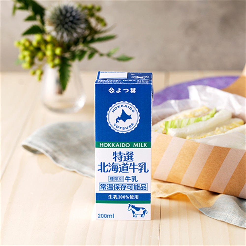 200ml carton of よつ葉特選北海道牛乳 (Hokkaido milk) standing on a table next to a sandwich.