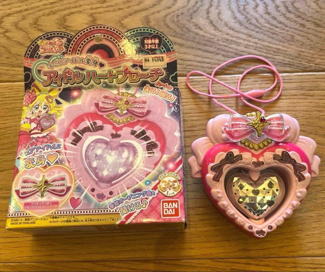 A pink heart-shaped Idol Heart Brooch toy displayed next to its original boxed packaging on a wooden surface.