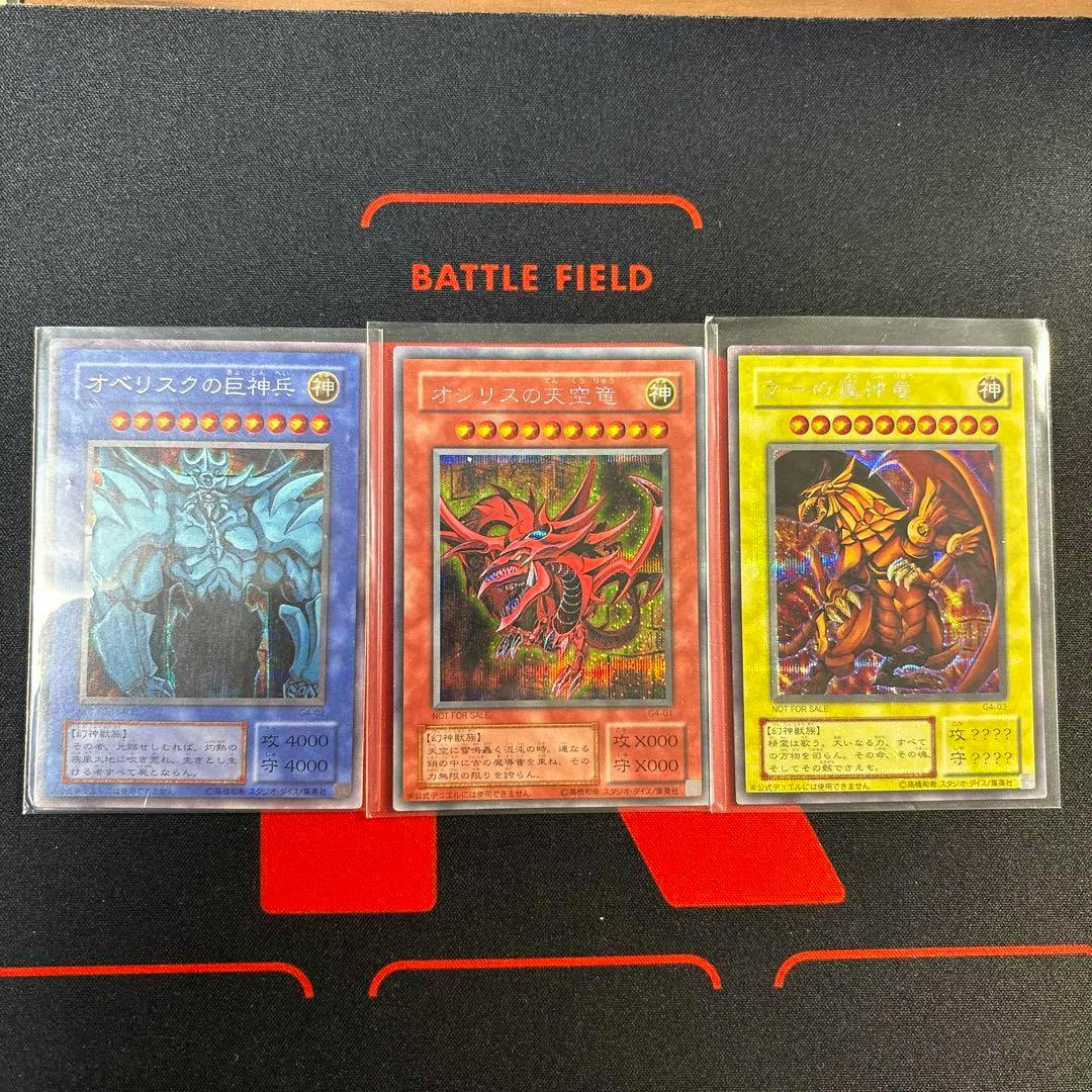 Three Yu-Gi-Oh trading cards (the three Egyptian God cards) in protective sleeves displayed side-by-side on a playmat.