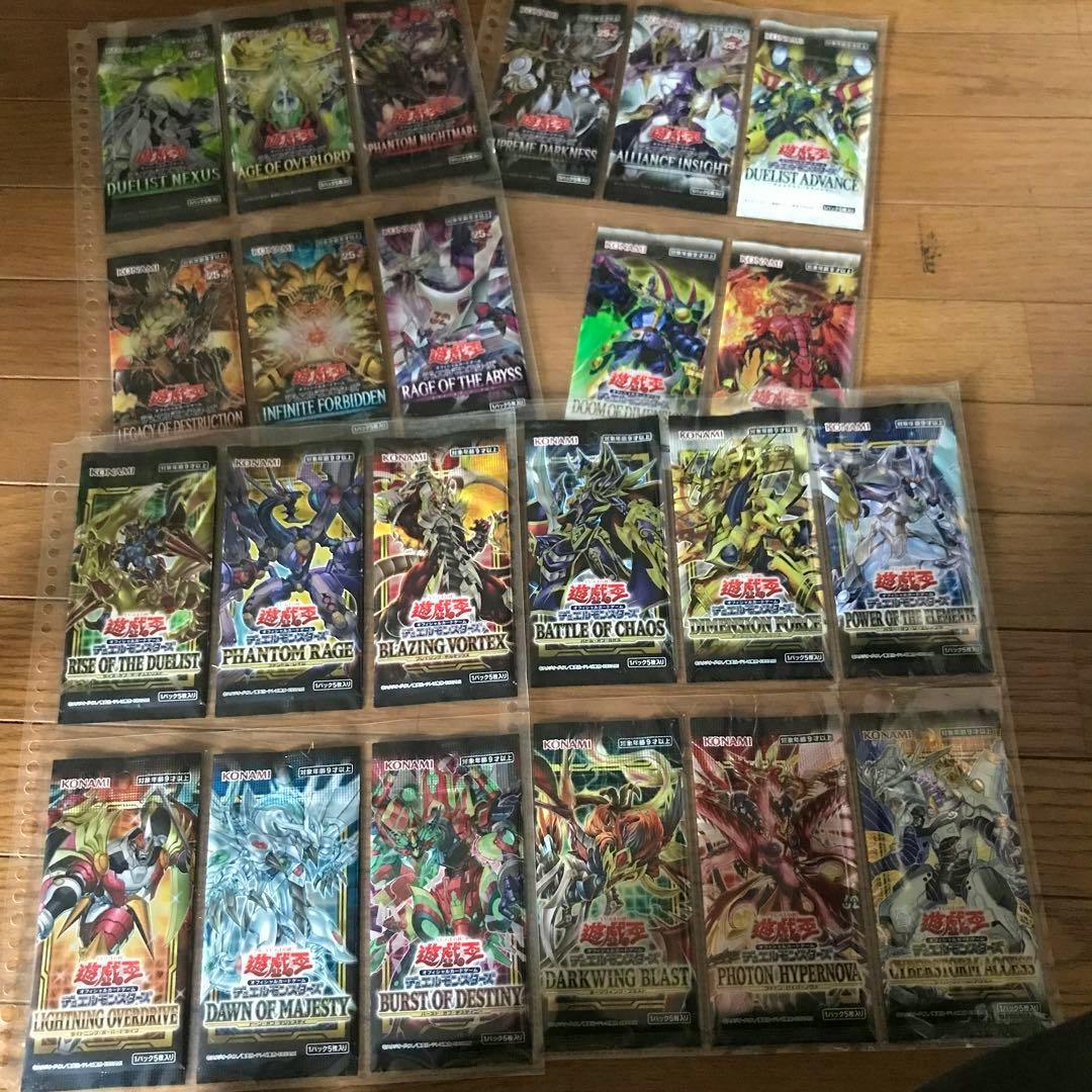 Photo of 23 unopened Yu-Gi-Oh booster packs arranged in plastic sleeves on a wooden floor.