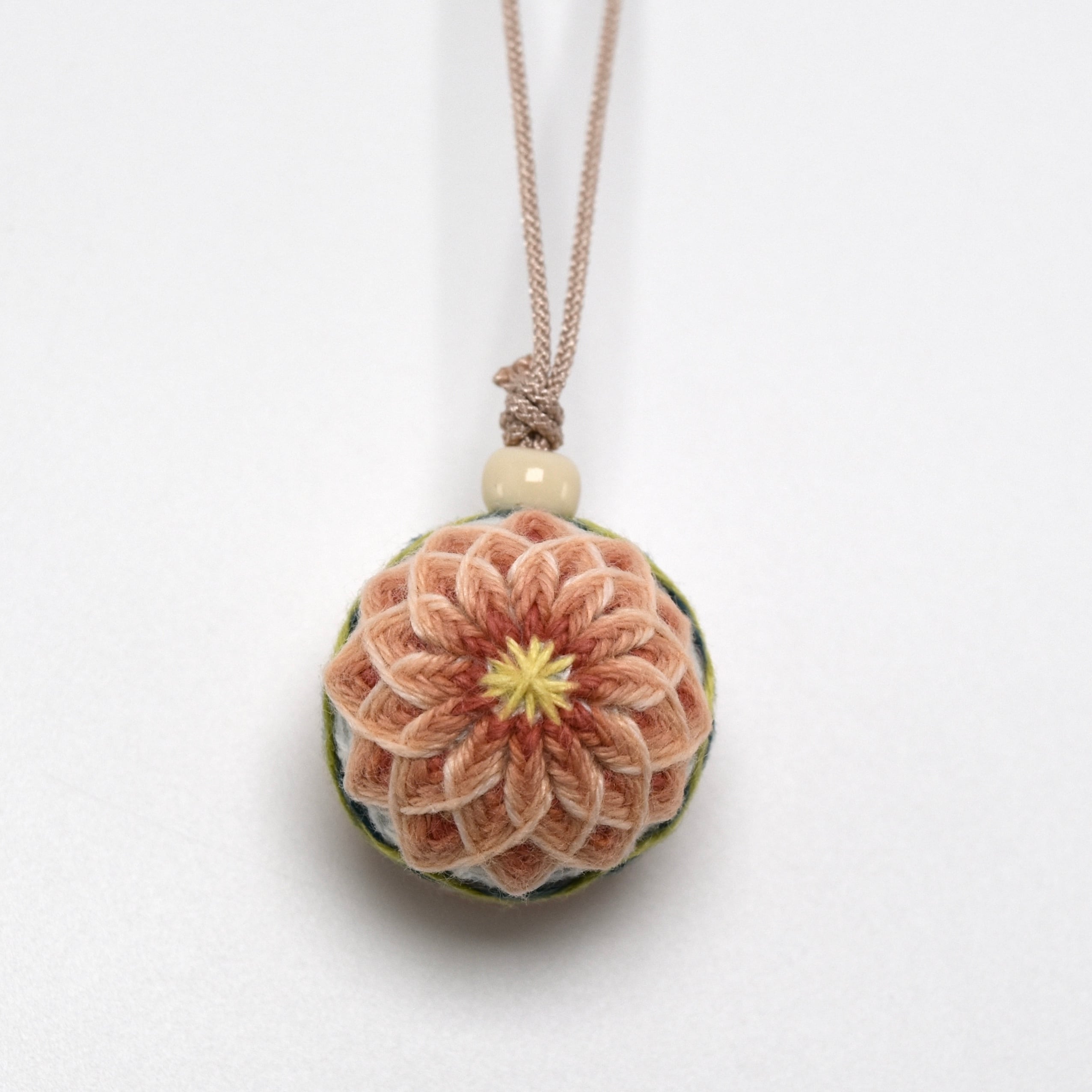 A fabric temari strap ornament with a pink layered embroidered flower design hanging from a beige cord with a bead on a white background.