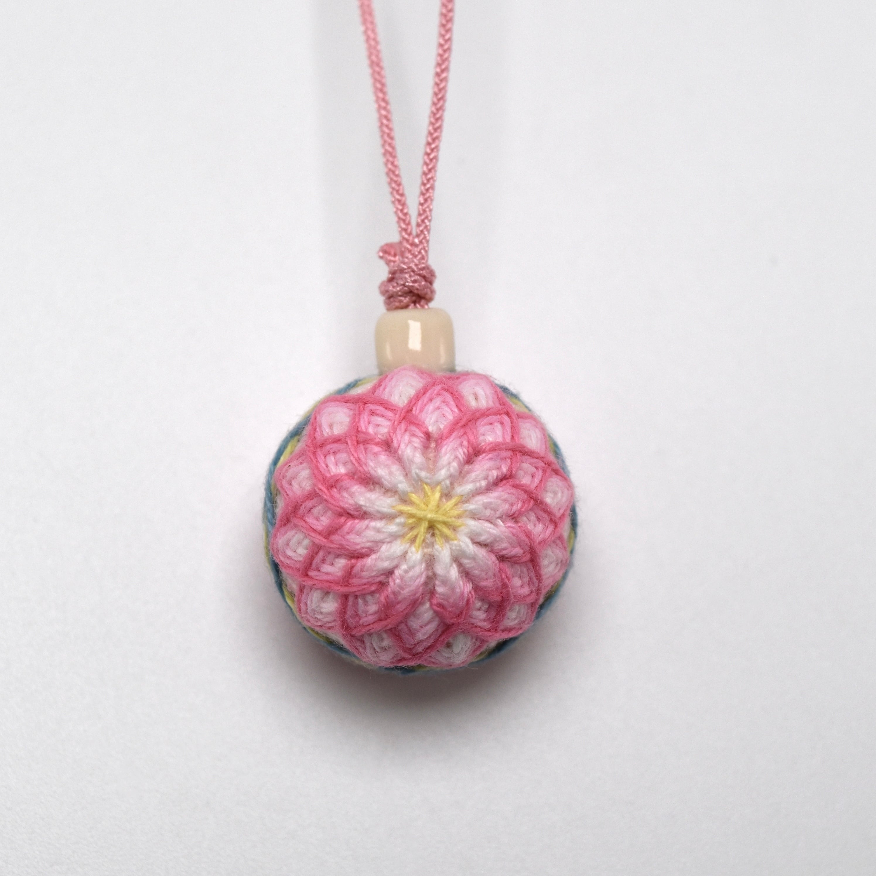A round pink and white embroidered temari strap ornament suspended from a pink cord against a white background.
