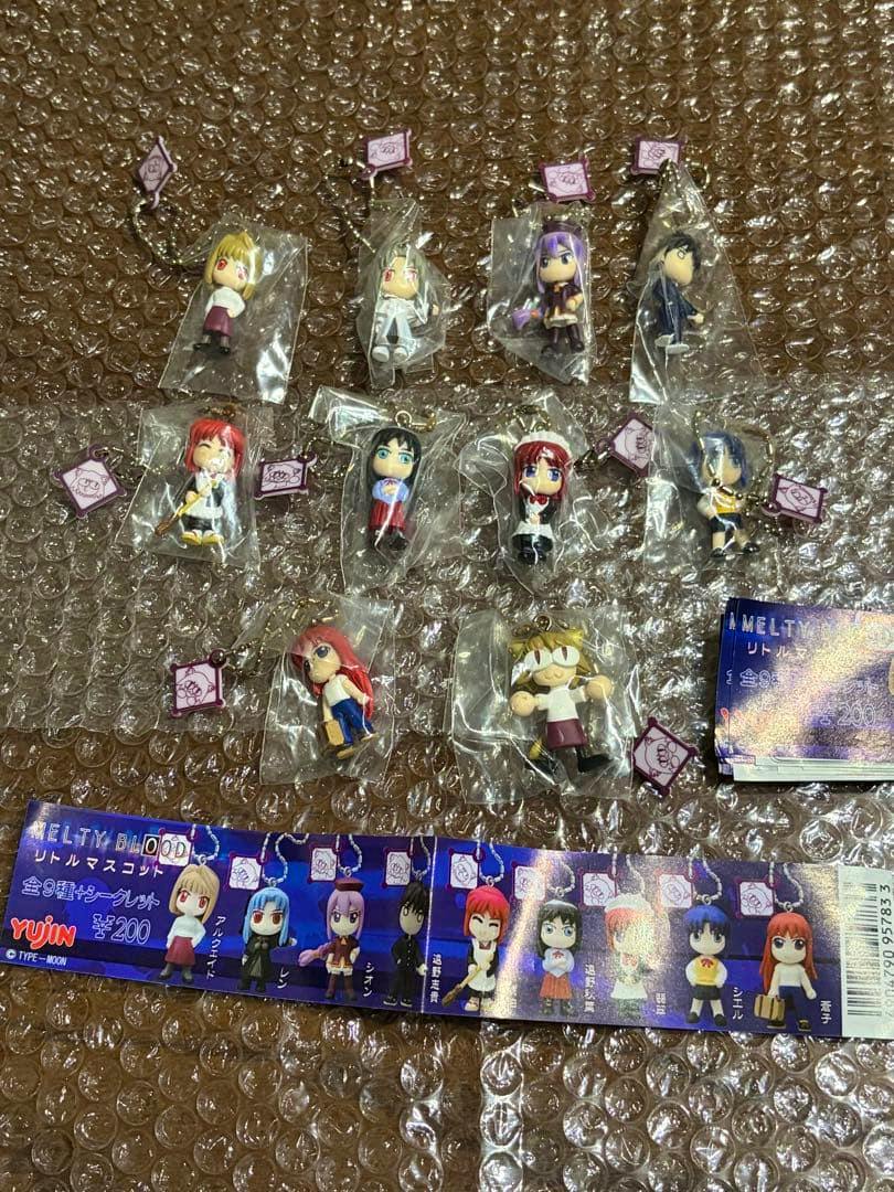 Photo of multiple YUJIN Melty Blood little mascot plastic keychain figures in individual bags arranged on bubble wrap with the character lineup strip visible.