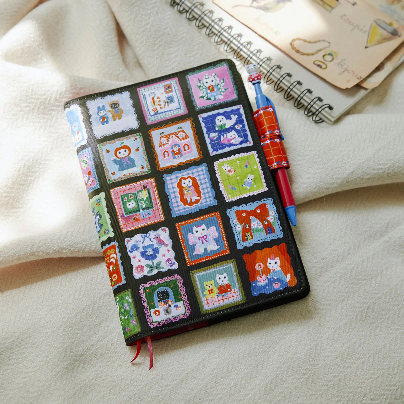 A polyester A5 cousin-size book cover showing a grid of colorful cat-themed square illustrations, photographed on a soft fabric with a pen attached.