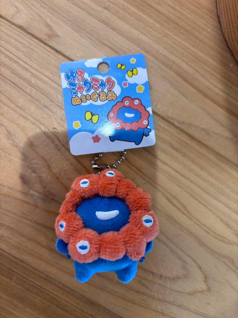 Small blue plush ball-chain keychain with orange ring and attached retail tag on a wooden surface.