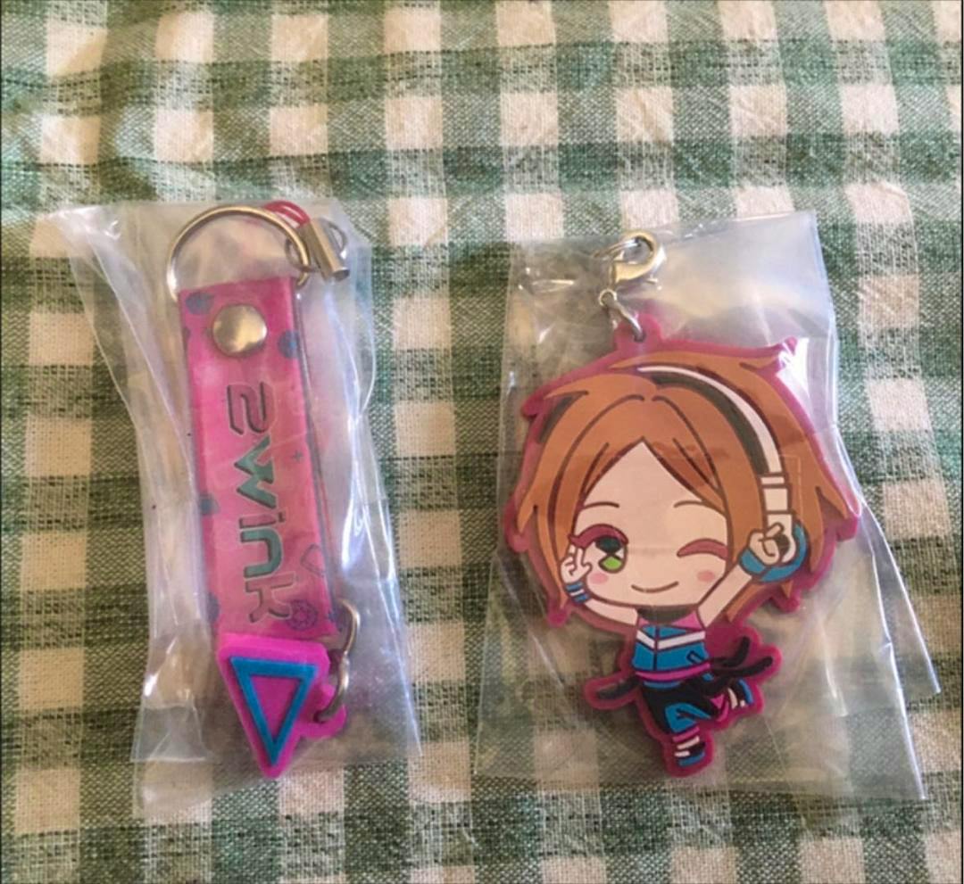 Two packaged rubber keychain items on a green-and-white checkered cloth: a pink strap with metal ring on the left and a chibi character rubber strap wearing headphones on the right.