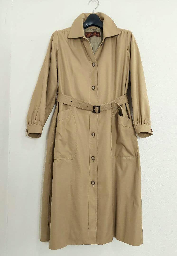 Beige long women's cotton coat with buttons and a matching belt hanging on a hanger against a white wall.