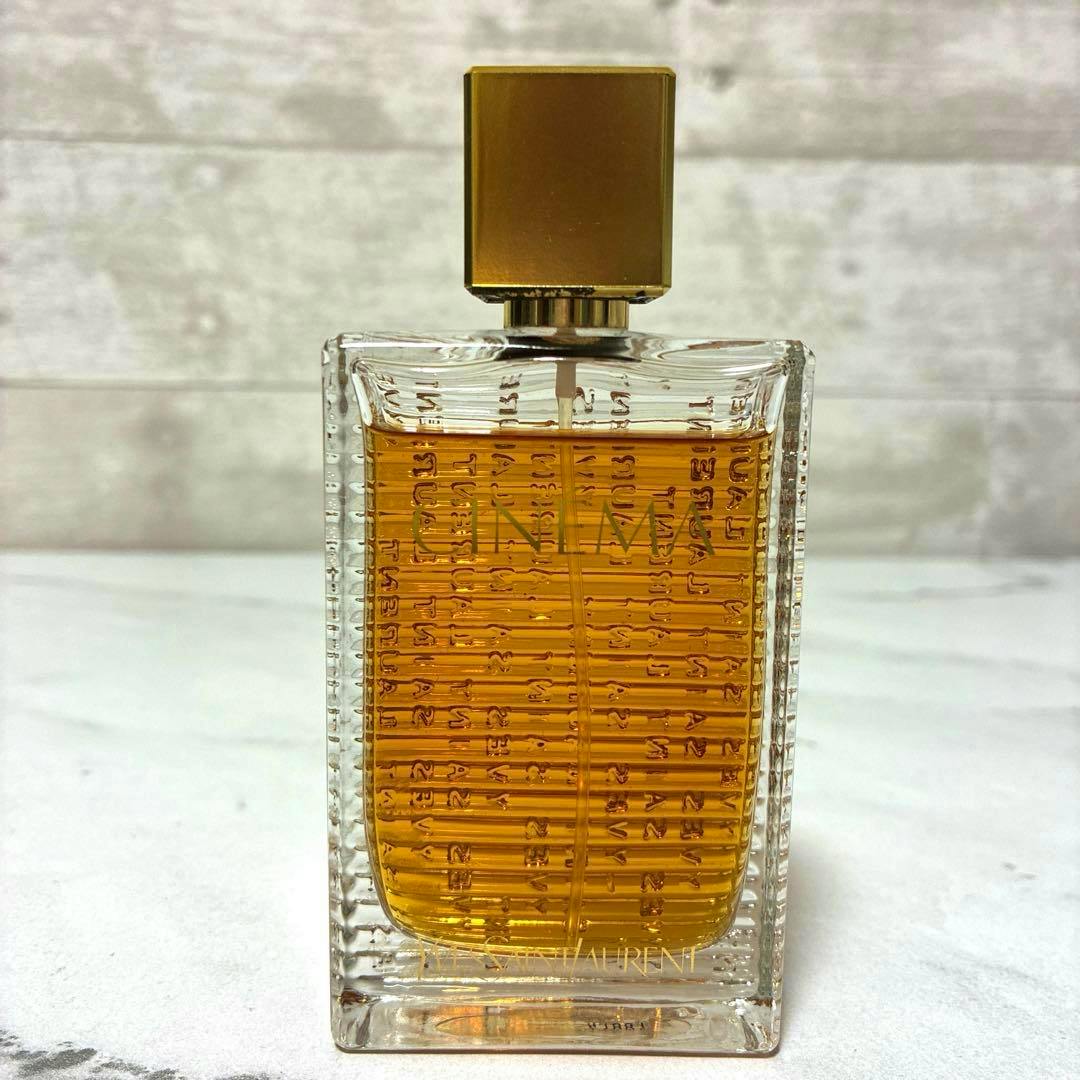 Clear rectangular glass bottle of Saint Laurent “Cinema” eau de toilette with amber liquid and a gold rectangular cap shown upright on a light surface.