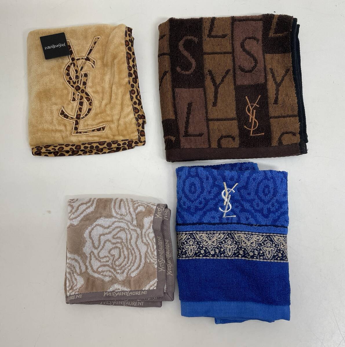Four folded Yves Saint Laurent hand towels in beige, brown, blue, and light brown are arranged on a white surface.