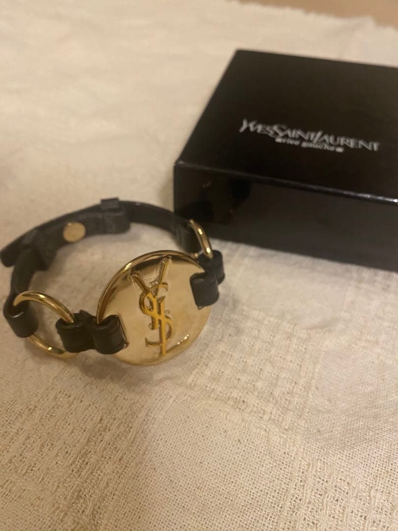 A gold Yves Saint Laurent round medallion cow-leather bracelet displayed on a light fabric surface with a black Yves Saint Laurent box behind it.