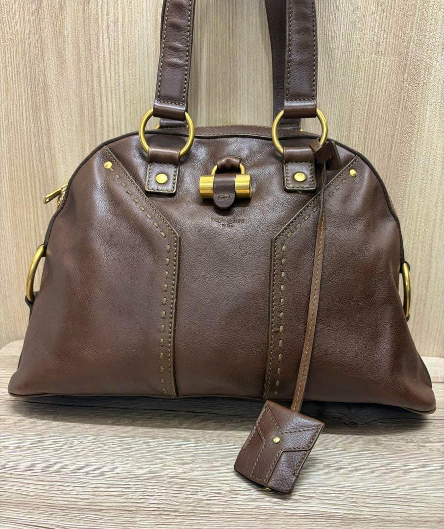 Brown leather Yves Saint Laurent Muse handbag shown front-facing on a wood surface with gold hardware and a hanging leather strap.