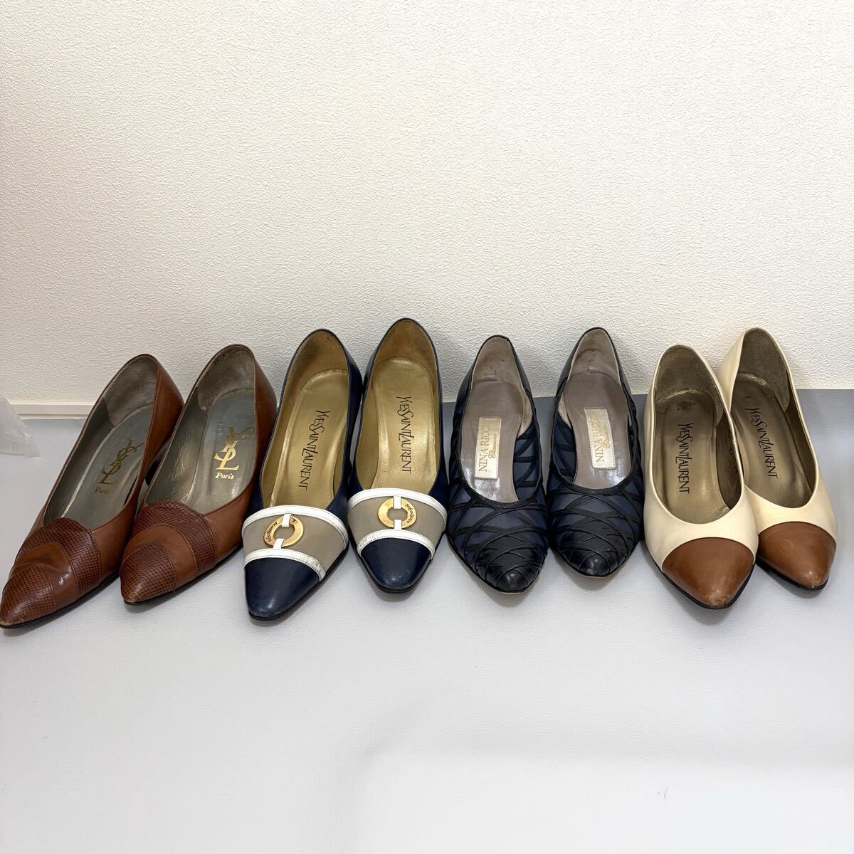 Several pairs of women's pumps arranged in a row on a light floor against a white wall, showing different colors and toe styles.