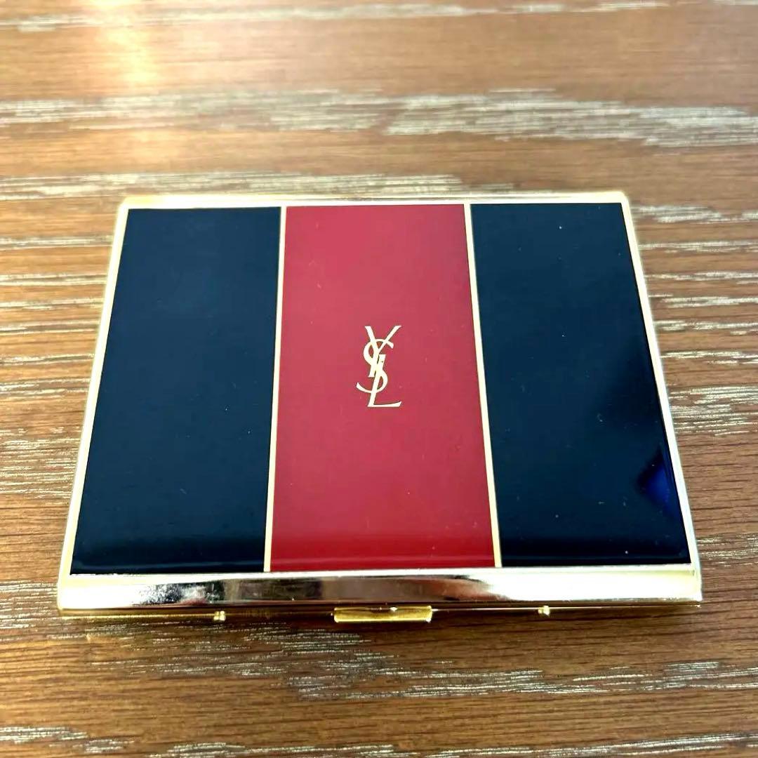 Rectangular YSL cigarette case with gold trim, black and red panels and a central YSL logo resting on a wood surface.