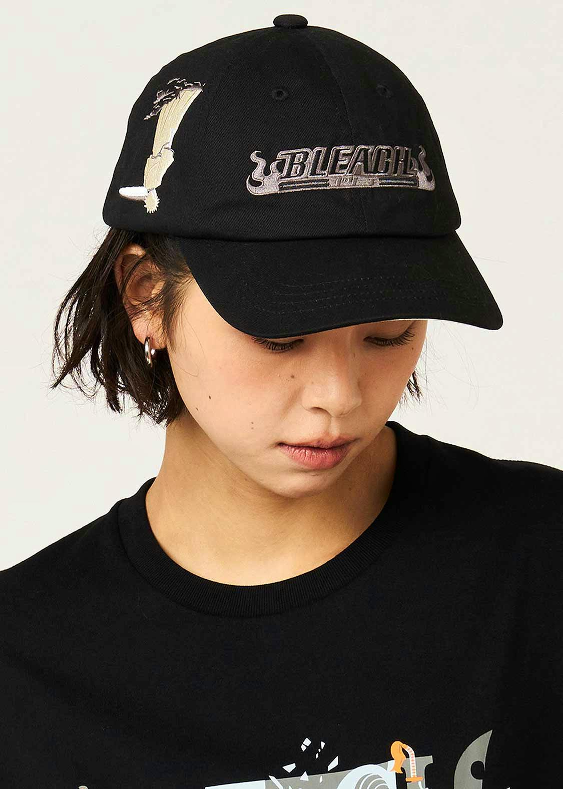 Front view of a black cotton baseball cap worn by a model, showing embroidered front graphics including the word "BLEACH".