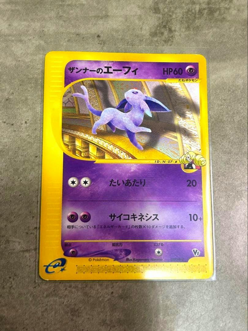 Front of the trading card ザンナーのエーフィ in a clear sleeve showing the card artwork and Japanese text on a gray surface.