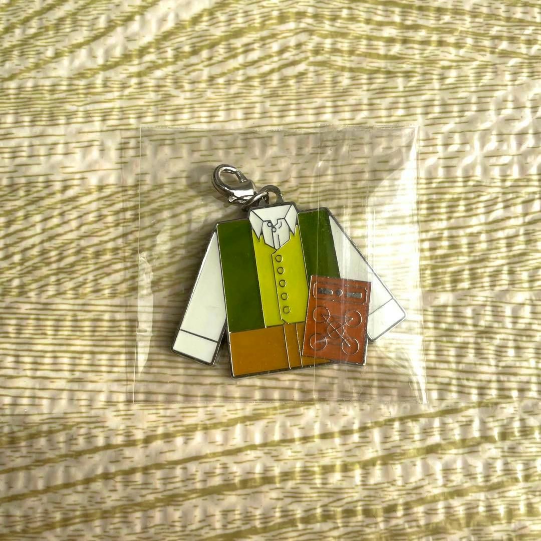 Front view of the zinc alloy bag charm showing a jacket-shaped enamel design in green and yellow with a small brown book, inside a clear plastic bag on textured fabric.