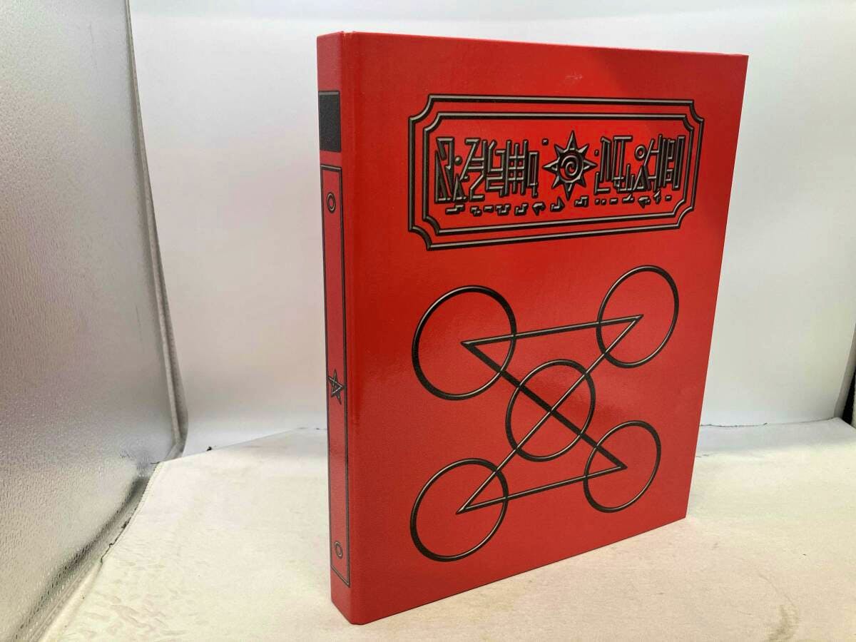 A red plastic binder with black circular-arrow symbols and a rectangular emblem, photographed standing on a white surface.