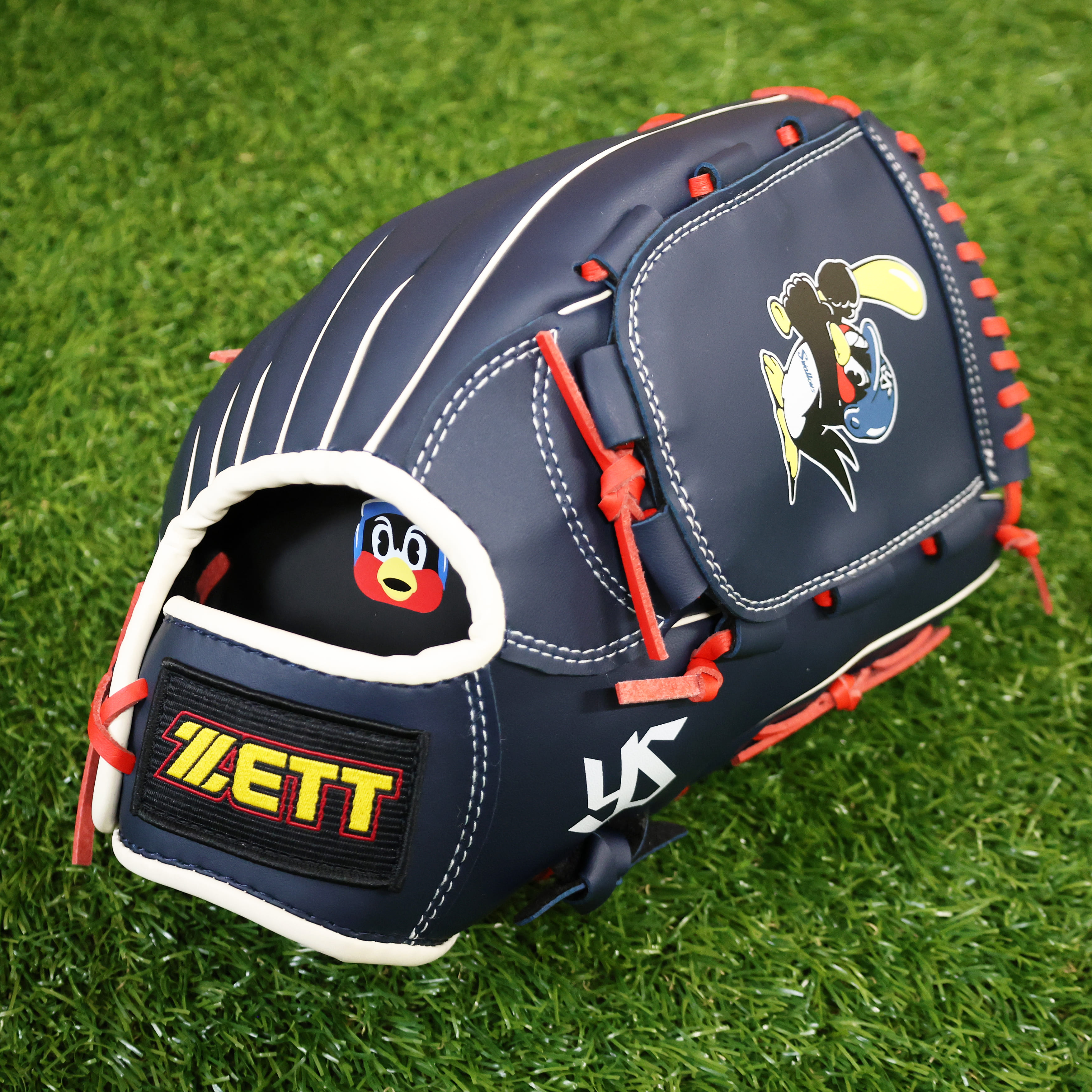 A navy ZETT soft baseball glove for adults with red lacing and a cartoon logo, shown on grass.