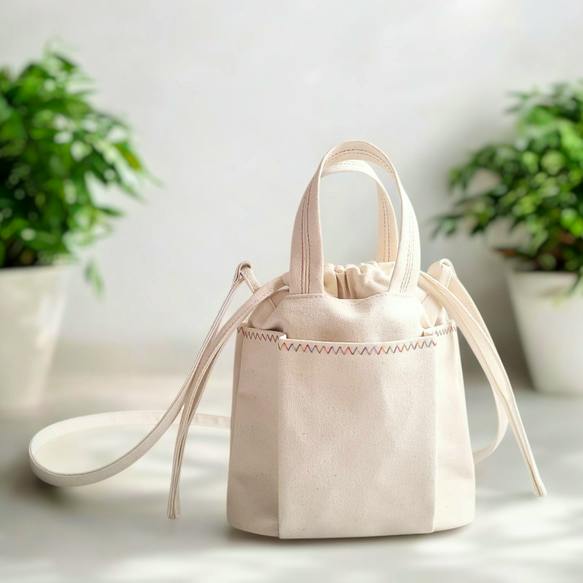 White cotton drawstring pochette bag with zigzag top stitching, short handles, and a long shoulder strap on a surface.