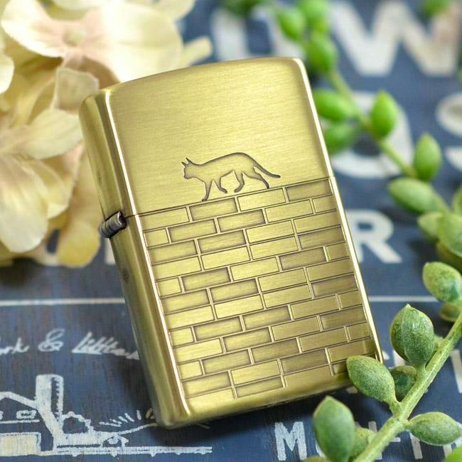 Brass-finish metal Zippo lighter case (insert removed) with an engraved cat walking above a brick-pattern design, shown on a blue decorative surface with plants.