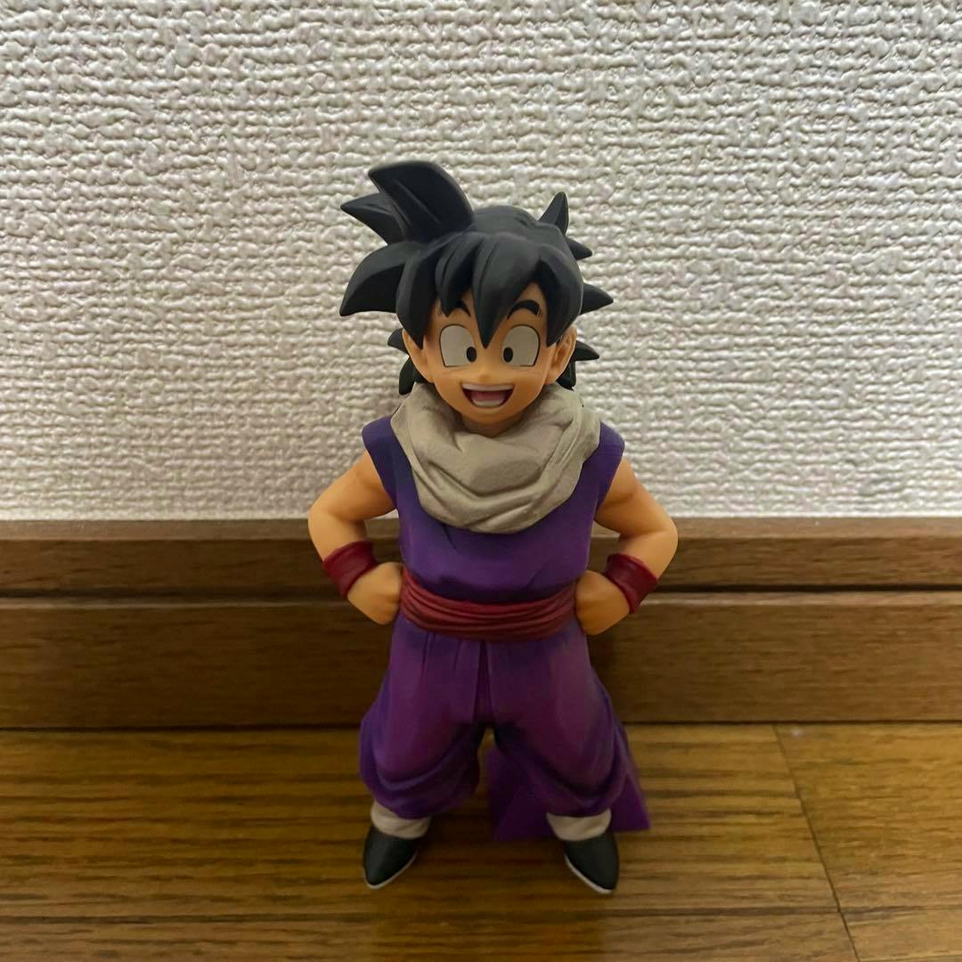 Front view of a plastic figure of Son Gohan wearing a purple outfit and beige scarf, standing on a wooden floor against a textured white wall.