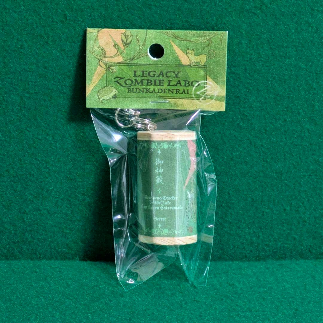 A green cylindrical omikuji keyholder in clear plastic packaging with a green header card, photographed against a green background.